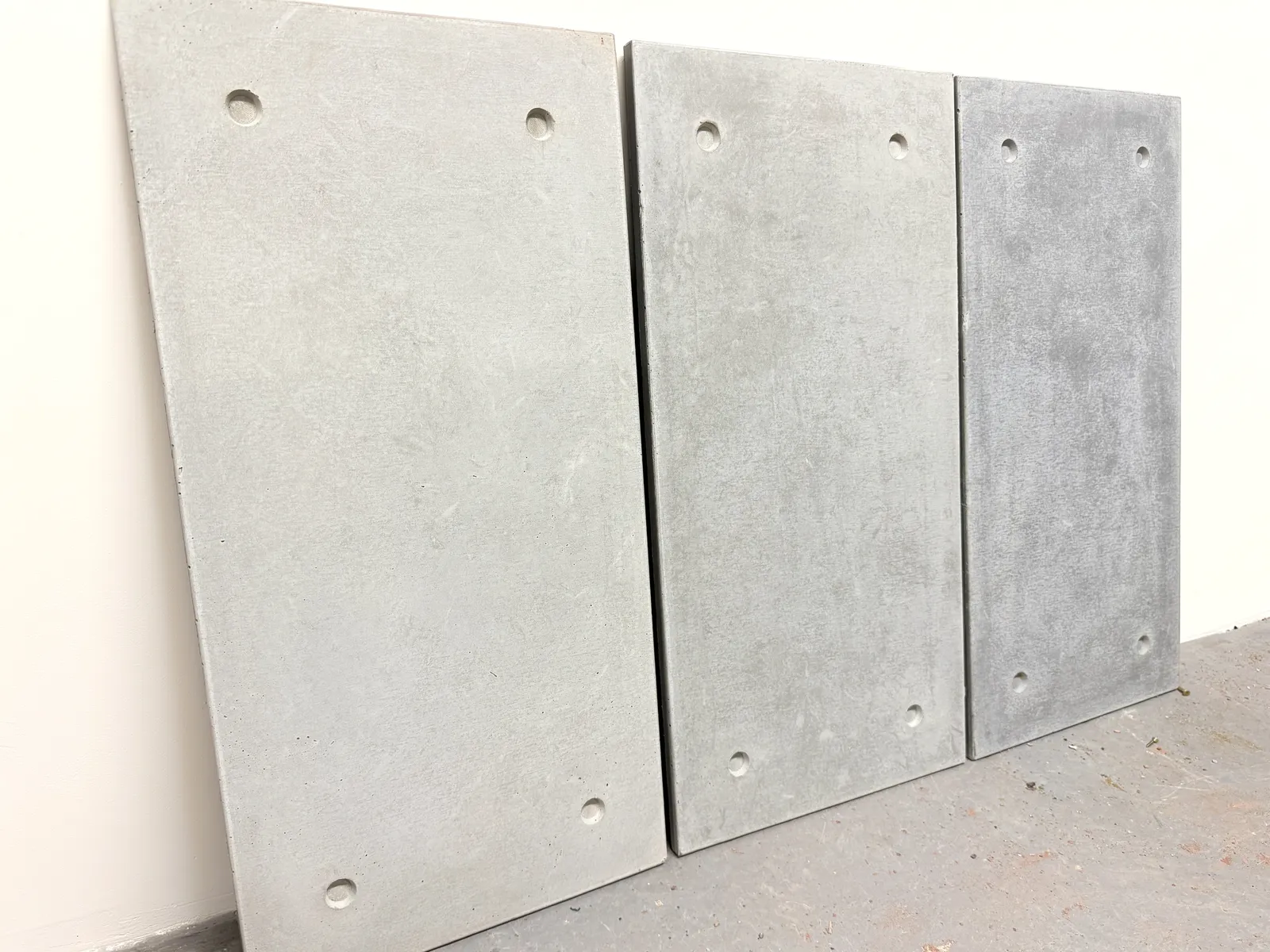 Three ROKA concrete cladding sample tiles in light, mid and dark grey leaning against a white wall
