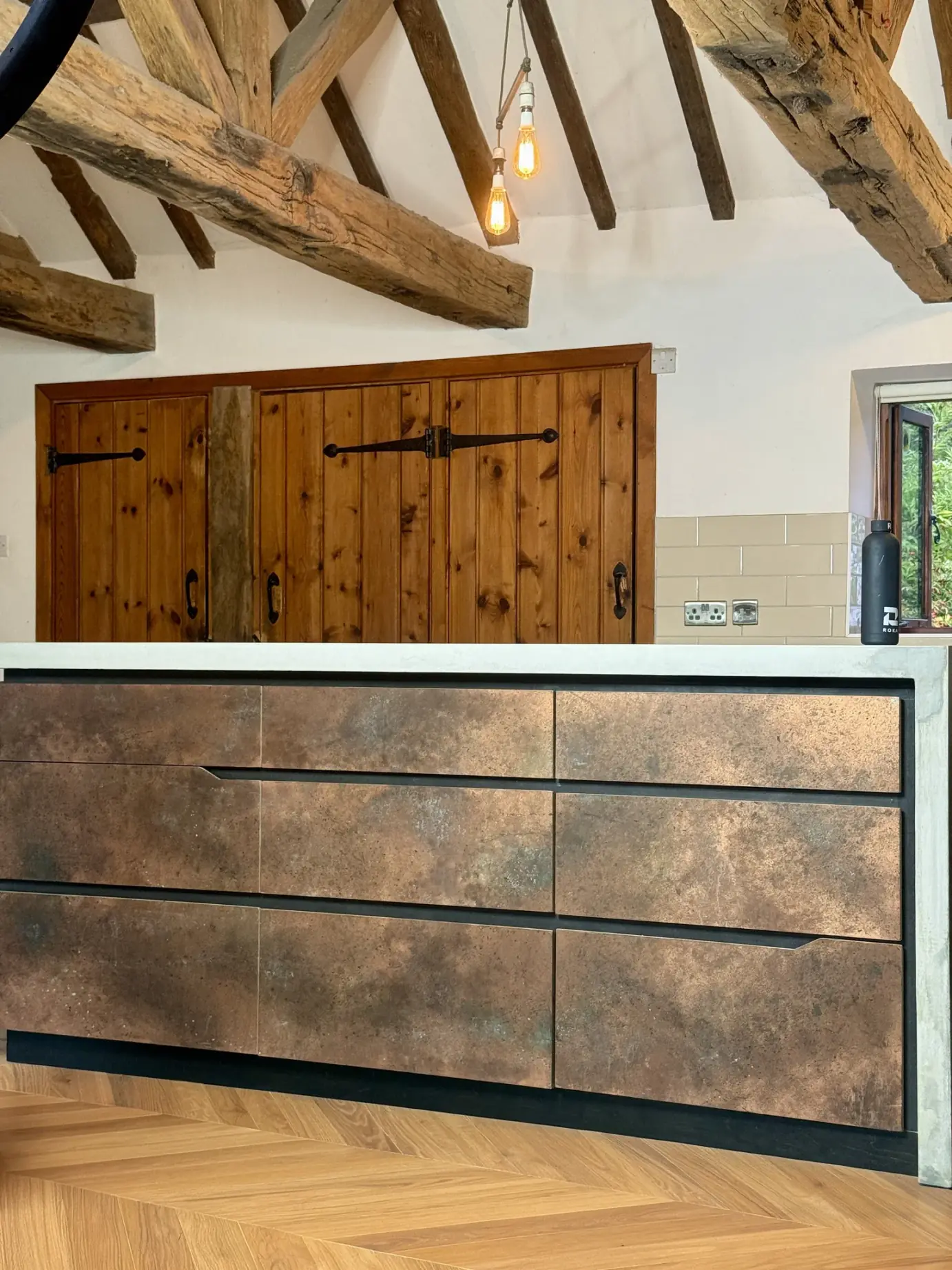 Kitchen island with copper patina drawer fronts and a pale concrete top with a waterfall end, oak herringbone floor and beamed ceiling