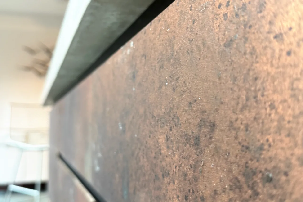 Close view of a copper-finish drawer front, mottled brown and gold, under the dark shadow gap and the concrete edge