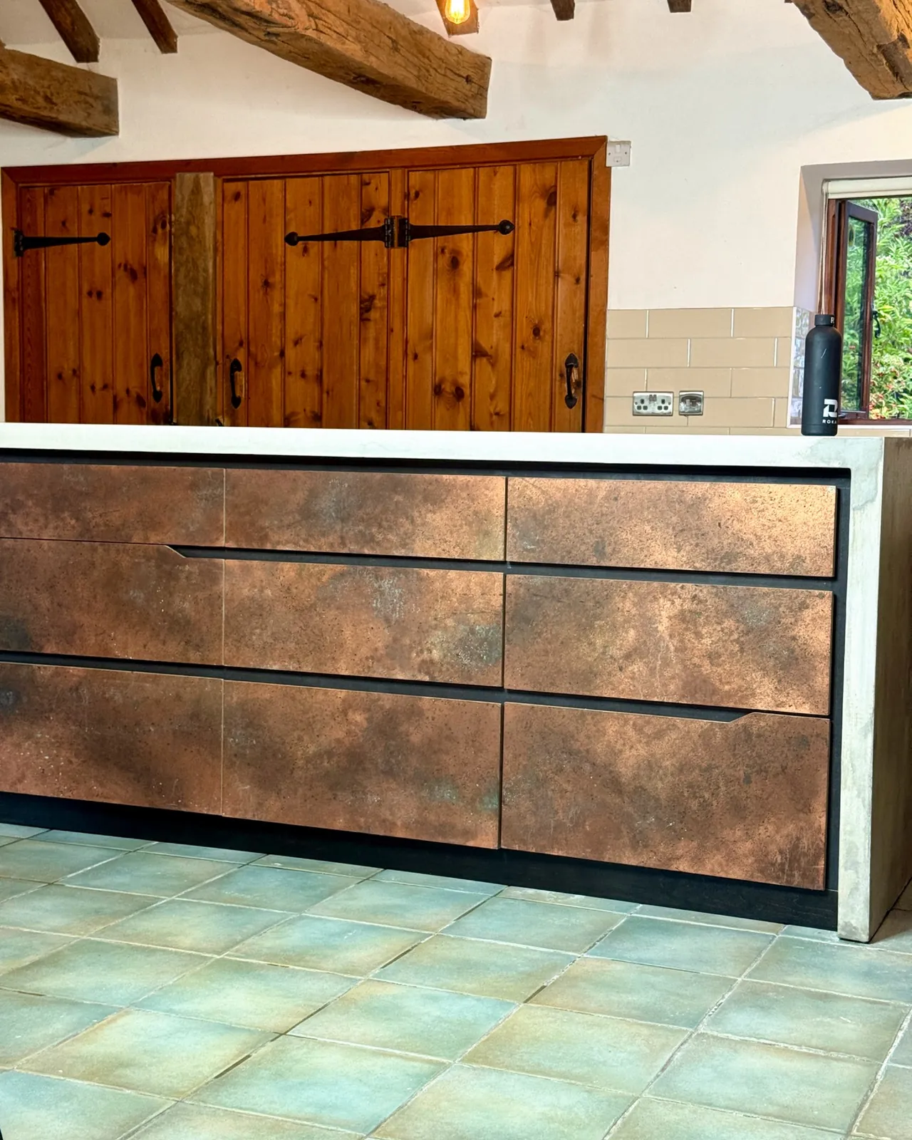 Long side of the concrete island, with nine copper-finish drawer fronts in a black frame and the concrete waterfall end running to the tiled floor