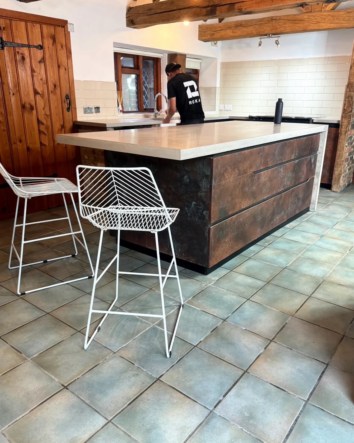 Pale concrete waterfall island with copper-finish drawer fronts and two white wire stools, in a barn kitchen with exposed beams